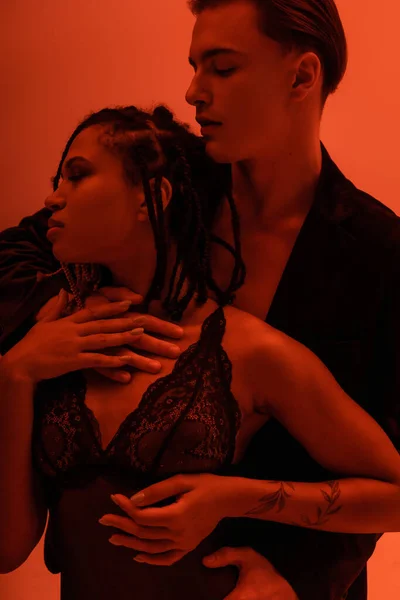 Joven hombre guapo en blazer negro abrazando encantadora mujer afroamericana con rastas y brazo tatuado, usando traje de encaje sobre fondo naranja con efecto de iluminación roja - foto de stock