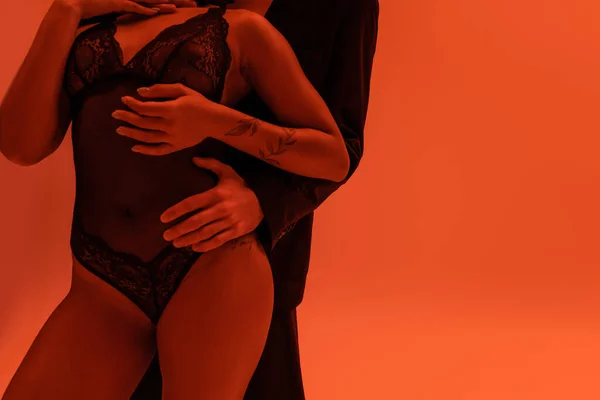 Частичный вид молодого человека в черном пиджаке, обнимающего сексуальную и страстную африканскую женщину с татуированной рукой в кружевном костюме на оранжевом фоне с эффектом освещения — Stock Photo