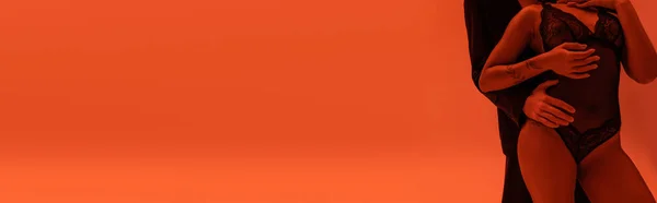 Teilaufnahme eines jungen Mannes im schwarzen Blazer, der eine Afroamerikanerin mit tätowiertem Arm umarmt und einen Spitzenbody auf orangefarbenem Hintergrund mit rotem Lichteffekt trägt, Banner — Stockfoto