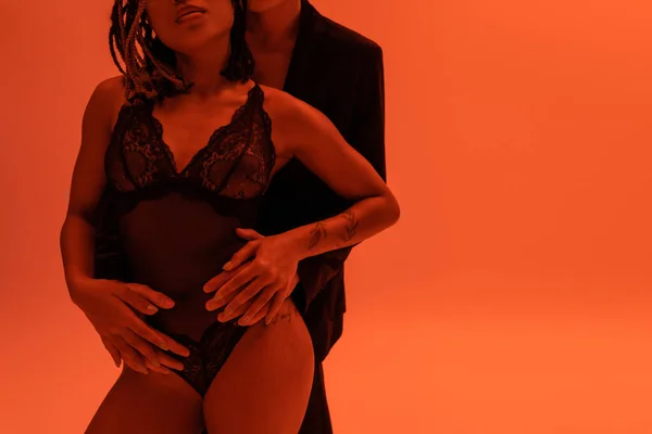 Cropped view of young man in blazer embracing seductive and sexy african american woman with tattooed arm, in black lace bodysuit on orange background with red lighting effect — Stock Photo