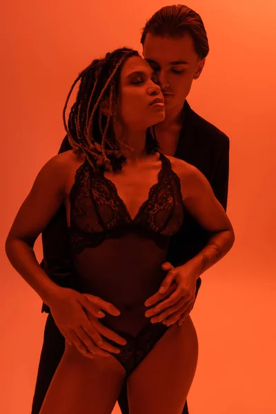 Young self-assured man seducing sexy african american woman with dreadlocks and closed eyes, wearing black lace bodysuit on orange background with red lighting effect — Stock Photo