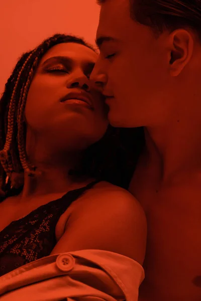 Young, shirtless and sexy man seducing and kissing passionate african american woman with dreadlocks and closed eyes, wearing black lace lingerie on orange background with red lighting effect — Stock Photo