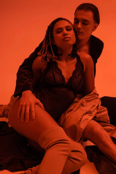 Уверенный мужчина в блейзере соблазняет африканскую американку в черном кружеве и обнимает ее ногу, сидя на оранжевом фоне с эффектом красного освещения — стоковое фото