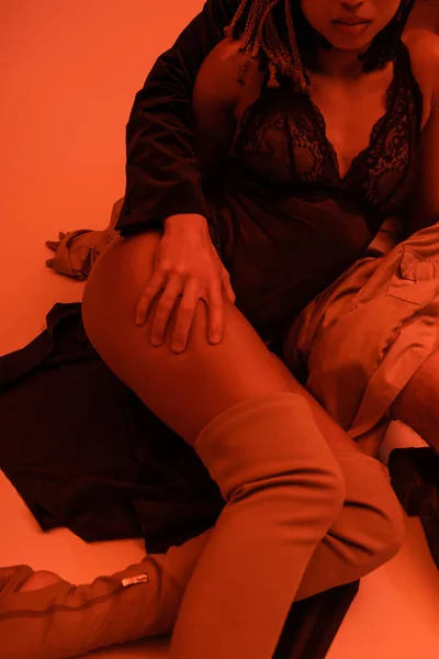 Partial view of young man embracing leg of sexy african american woman in over knee boots and black lace bodysuit sitting on clothes on orange background with red lighting effect — Stock Photo