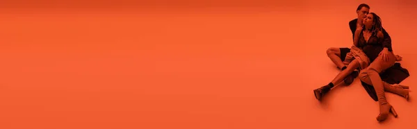 Hochwinkelaufnahme von sexy interrassischen Paar, junger schöner Mann und afrikanisch-amerikanische Frau in schwarzem Spitzenbody und Overknee-Stiefeln sitzt auf Kleidung auf orangefarbenem Hintergrund mit roter Beleuchtung, Banner — Stockfoto