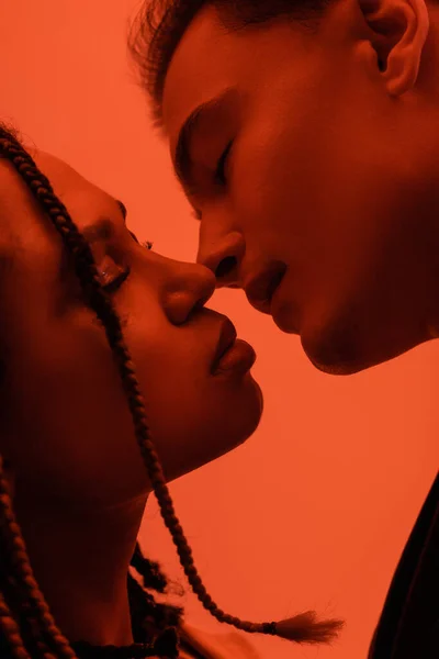 Close up view of young and sexy interracial couple in love, young man and african american woman with dreadlocks kissing with closed eyes on orange background with red lighting effect — Stock Photo
