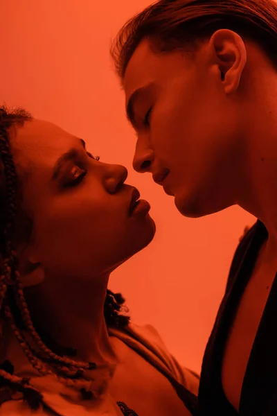 Side view of young and sensual african american woman with dreadlocks and handsome man with closed eyes kissing on orange background with red lighting effect — Stock Photo