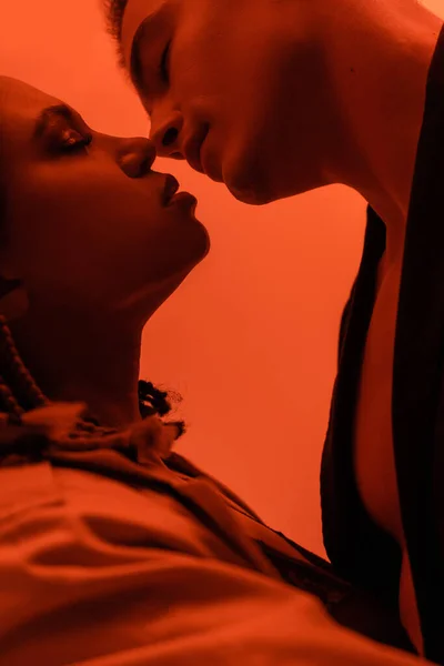 Vista lateral de joven y romántica pareja interracial enamorada, hombre carismático y sensual mujer afroamericana besándose con los ojos cerrados sobre fondo naranja con efecto de luz roja - foto de stock