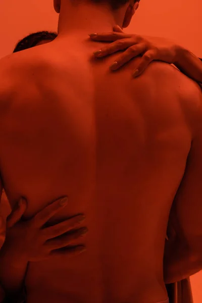 Momento intimo di giovane e senza maglietta uomo vicino appassionato donna afroamericana abbracciando la sua schiena muscolare su sfondo arancione con effetto luce rossa — Foto stock