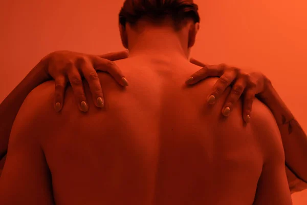 Young and shirtless man with muscular back near passionate african american woman embracing his shoulders on orange background with red lighting effect — Stock Photo