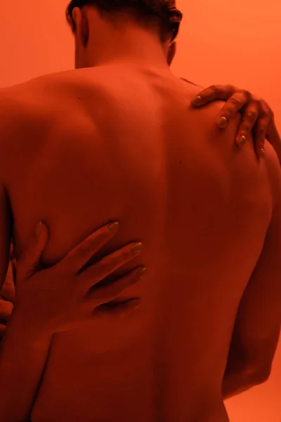 Rückansicht des jungen, hemdlosen und sexy Mannes in der Nähe einer leidenschaftlichen Afroamerikanerin, die seinen muskulösen Körper auf orangefarbenem Hintergrund mit rotem Lichteffekt umarmt — Stockfoto