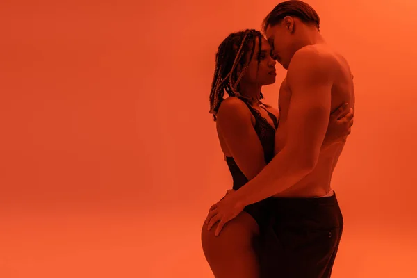 Vue latérale d'un jeune homme torse nu avec torse musclé embrassant une femme américaine africaine sexy avec dreadlocks, portant un body en dentelle noire sur fond orange avec un effet d'éclairage rouge — Photo de stock