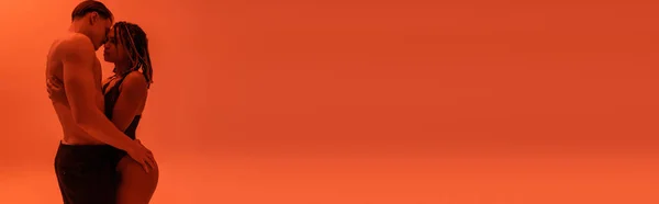 Vista laterale di giovane uomo senza camicia in pantaloni neri che abbraccia provocatoria donna afroamericana in pizzo body su sfondo arancione con effetto luce rossa, banner — Foto stock