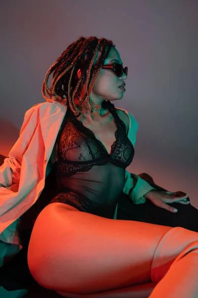 Seductive african american woman with dreadlocks wearing dark sunglasses, black lace bodysuit and beige trench coat sitting on huge tire and looking away on grey background with red lighting — Stock Photo