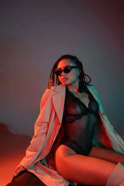 Expressivo e elegante modelo afro-americano em óculos escuros, macacão preto e casaco bege sentado em um pneu enorme e olhando para o fundo cinza com iluminação vermelha — Fotografia de Stock