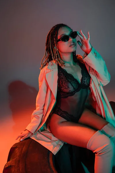 Mulher americana africana glamourosa e atrevida ajustando óculos escuros e elegantes enquanto sentado em um pneu enorme em um macacão preto e casaco bege em fundo cinza com iluminação vermelha — Fotografia de Stock