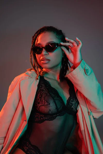 Young and sexy african american woman with dreadlocks adjusting dark sunglasses while posing in black lace bodysuit and beige trench coat on grey background with red lighting — Stock Photo
