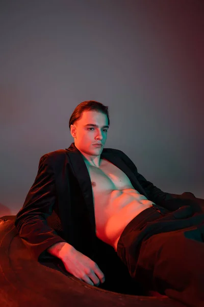 Self-assured and glamorous man wearing black stylish blazer on shirtless muscular body, sitting on huge tire and looking away on grey background with red lighting — Stock Photo