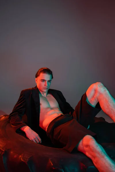 Провокационный и модный молодой человек в черных шортах и модный пиджак на мускулистом теле без рубашки сидит на огромной шине на сером фоне с красным освещением — стоковое фото