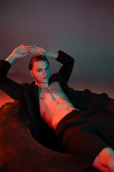 Confident and sexy man in black trendy blazer on shirtless muscular torso posing on huge tire with hands above head on grey background with red lighting — стоковое фото