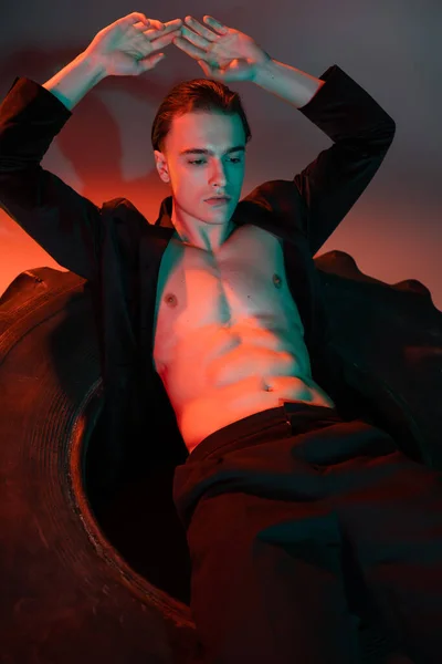 Junger und sexy Mann trägt schwarzen Blazer auf hemdlosem muskulösem Oberkörper, sitzt auf riesigen Reifen und posiert mit den Händen über dem Kopf auf grauem Hintergrund mit roter Beleuchtung — Stockfoto