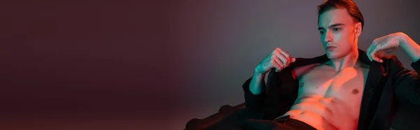 Hombre joven, elegante y sexy que usa chaqueta negra en el cuerpo muscular sin camisa mientras está sentado y posando sobre un enorme neumático sobre un fondo gris con iluminación roja, pancarta - foto de stock
