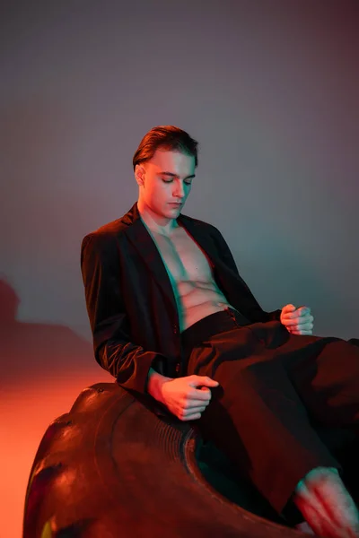 Joven, hombre guapo y sexy en blazer negro de moda en torso muscular sin camisa sentado y posando sobre un neumático enorme sobre fondo gris con iluminación roja - foto de stock