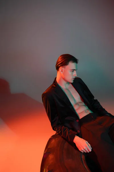 Homem jovem e sexy com cabelo morena, vestindo blazer preto no corpo muscular sem camisa, posando em pneu enorme enquanto sentado em fundo cinza com iluminação vermelha — Fotografia de Stock
