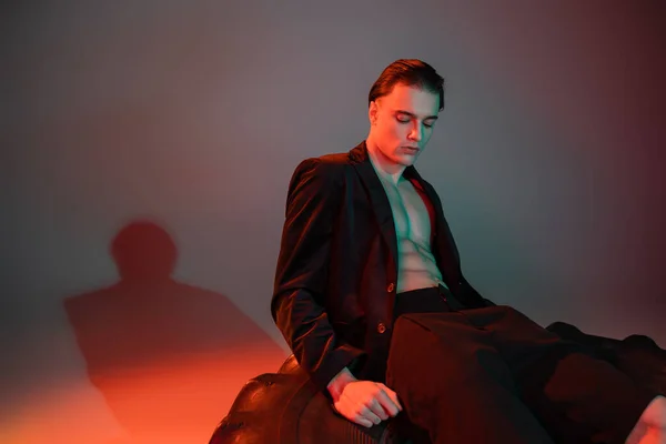 Junger, sexy und modischer Mann sitzt auf einem riesigen Reifen und posiert in schwarzen Shorts und stylischem Blazer auf hemdlosem muskulösem Körper auf grauem Hintergrund mit roter Beleuchtung — Stockfoto