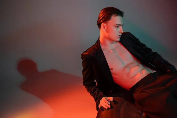 Young good looking man with brunette hair sitting on huge tire while posing in black blazer on shirtless muscular torso on grey background with red lighting — Stock Photo