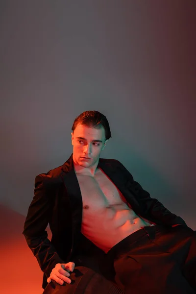 Young, handsome and sexy man in black stylish blazer on shirtless muscular body sitting on huge tire and looking away on grey background with red lighting — Stock Photo