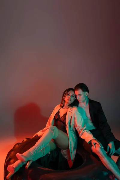 Модная межрасовая пара, сидящая на огромной шине, мужчина без рубашки в черном пиджаке и африканская американка в кружевном костюме, бежевом плаще и на коленных сапогах на сером фоне с красным освещением — стоковое фото