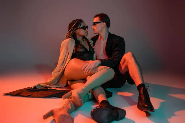 Сексуальная межрасовая пара в солнечных очках, африканская американка в черном кружевном костюме, бежевый плащ и ботинки на коленях, уверенный мужчина в блейзере на сером фоне с красным освещением — стоковое фото