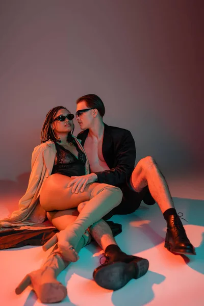 Full length of stylish man in dark sunglasses and black blazer embracing leg of african american woman in lace bodysuit, beige trench coat and over knee boots on grey background with red lighting — Stock Photo