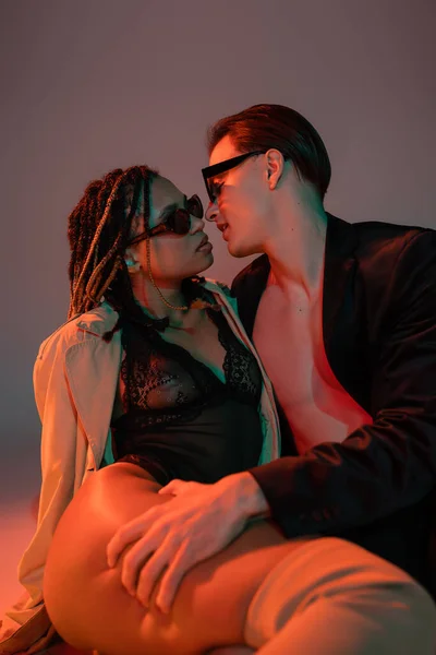 Young and confident man in dark sunglasses and black blazer seducing provocative african american man in lace bodysuit and beige trench coat on grey background with red lighting — Stock Photo