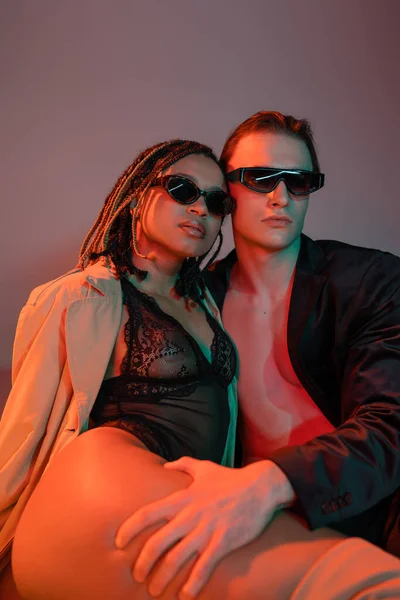 Sexy y de moda pareja interracial en gafas de sol oscuras, hombre joven en chaqueta negra y mujer afroamericana en traje de encaje y gabardina beige sobre fondo gris con iluminación roja - foto de stock