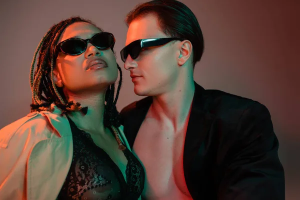 Сексуальная межрасовая пара в темных солнцезащитных очках, харизматичный мужчина в черном пиджаке и африканская американка в кружевном белье и бежевом плаще на сером фоне с красным освещением — стоковое фото
