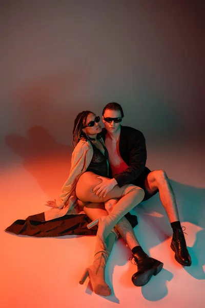 High angle view of trendy man in black blazer and shorts near african american woman in dark sunglasses, lace bodysuit, beige trench coat and over knee boots on grey background with red lighting — Stock Photo