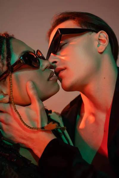 Momento íntimo de sexy pareja interracial besándose en gafas de sol oscuras, mujer afroamericana con rastas y hombre joven en chaqueta negra sobre fondo gris con iluminación roja - foto de stock