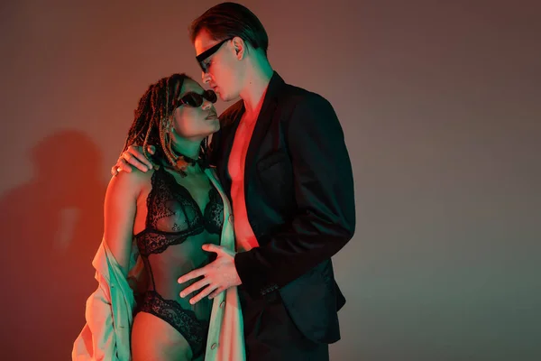 Glamorous man in dark sunglasses and blazer embracing provocative african american woman in black lace bodysuit and beige trench coat on grey background with red lighting — Stock Photo