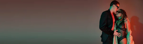 Jeune homme confiant en lunettes de soleil sombres et blazer noir embrassant femme afro-américaine en costume de dentelle et trench coat beige sur fond gris avec éclairage rouge, bannière — Photo de stock