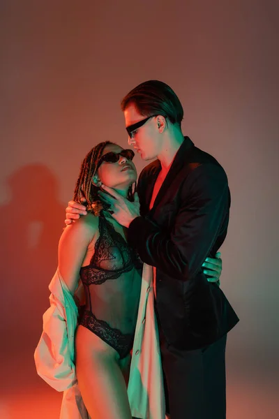 Young and confident man in dark sunglasses and black blazer embracing tempting african american woman in lace bodysuit and beige trench coat on grey background with red lighting — Stock Photo