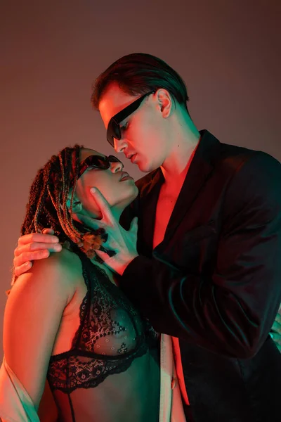 Homem jovem e auto-confiante em óculos escuros e blazer preto seduzindo encantadora mulher afro-americana em terno de renda em fundo cinza com iluminação vermelha — Fotografia de Stock
