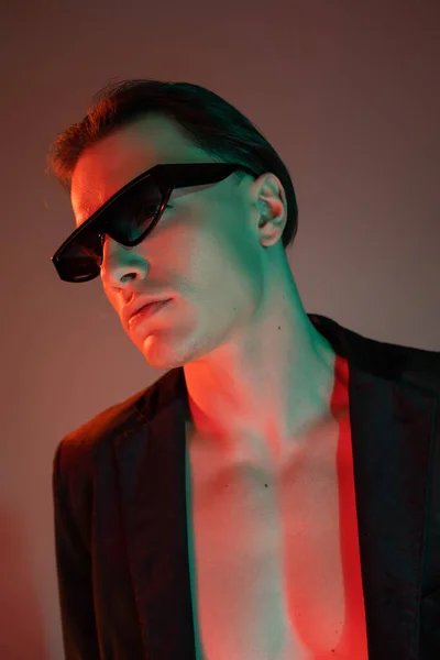 Retrato del joven hombre de pecho desnudo en chaqueta negra de moda y gafas de sol oscuras con estilo mirando hacia otro lado mientras posa sobre fondo gris con iluminación roja - foto de stock