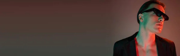 Retrato de glamuroso hombre desnudo con el pelo moreno posando en chaqueta negra y gafas de sol oscuras de moda sobre fondo gris con iluminación roja y espacio para copiar, pancarta - foto de stock