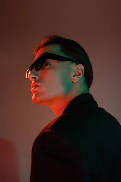 Homem jovem e carismático com cabelo morena posando em óculos escuros de sol e blazer preto enquanto olha para o fundo cinza com iluminação vermelha — Fotografia de Stock