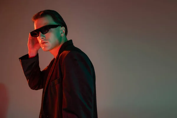 Young, fashionable and confident man adjusting dark stylish sunglasses while standing and posing in black blazer on grey background with red lighting and copy space — Stock Photo