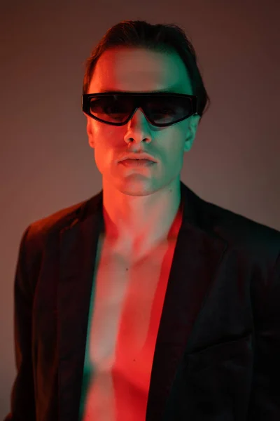 Portrait of young and fashionable man in dark sunglasses and stylish blazer looking at camera while standing and posing on grey background with red lighting — Stock Photo
