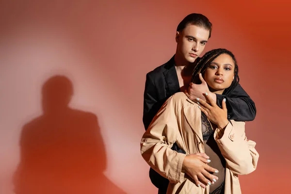 Sexy gemischtrassiges Paar, stylischer Mann im schwarzen Blazer und afrikanisch-amerikanische Frau im Spitzenbody und beigem Trenchcoat, die in die Kamera auf rotem und pinkem Hintergrund blickt — Stockfoto