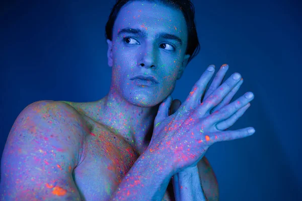 Jeune, homme charismatique et torse nu avec corps musclé en peinture au néon colorée posant avec les mains jointes et regardant loin sur fond bleu avec effet d'éclairage cyan — Photo de stock